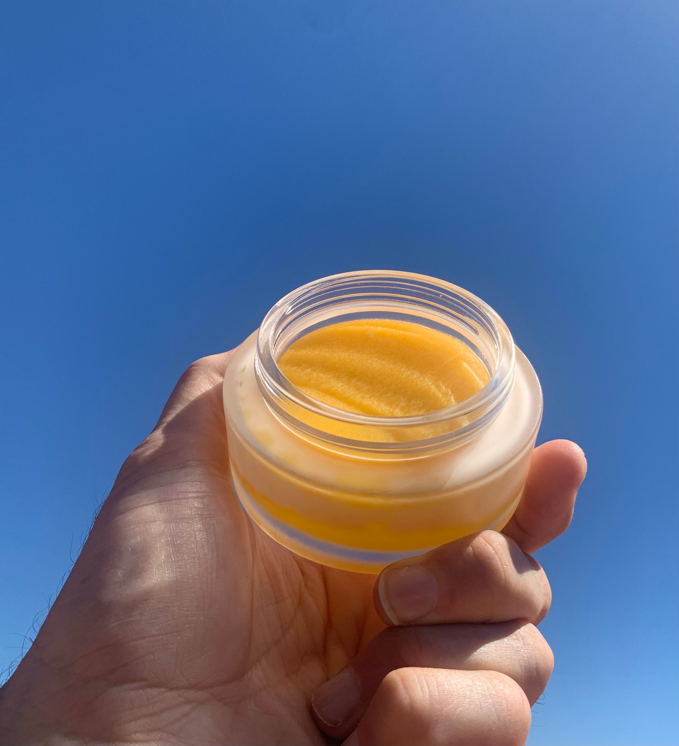 Hand holding a glass jar of yellow cream against a clear blue sky