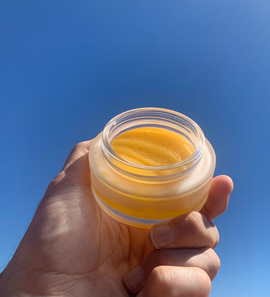 Hand holding a glass jar of yellow cream against a clear blue sky