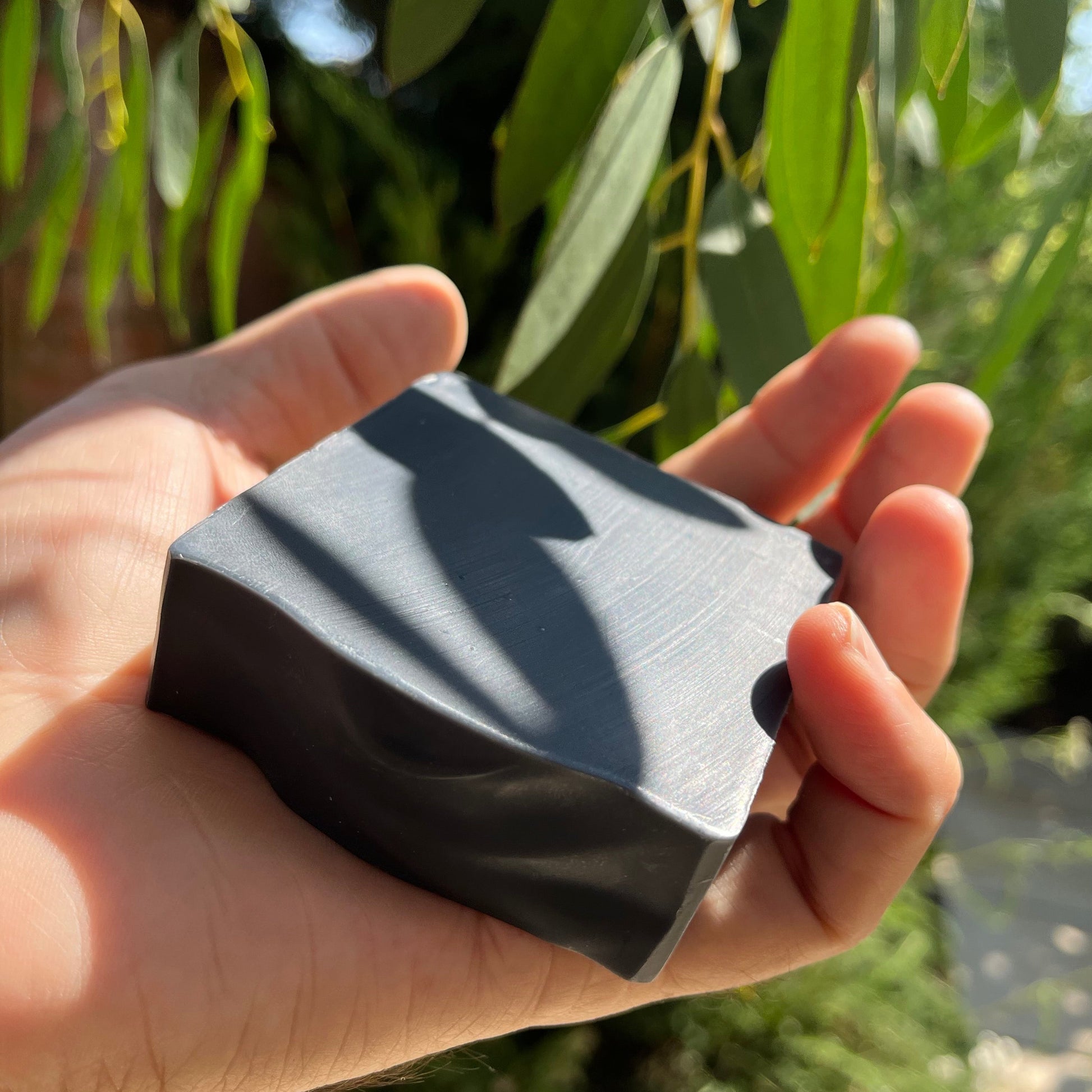 Hand holding a black bar of soap with green foliage in the background