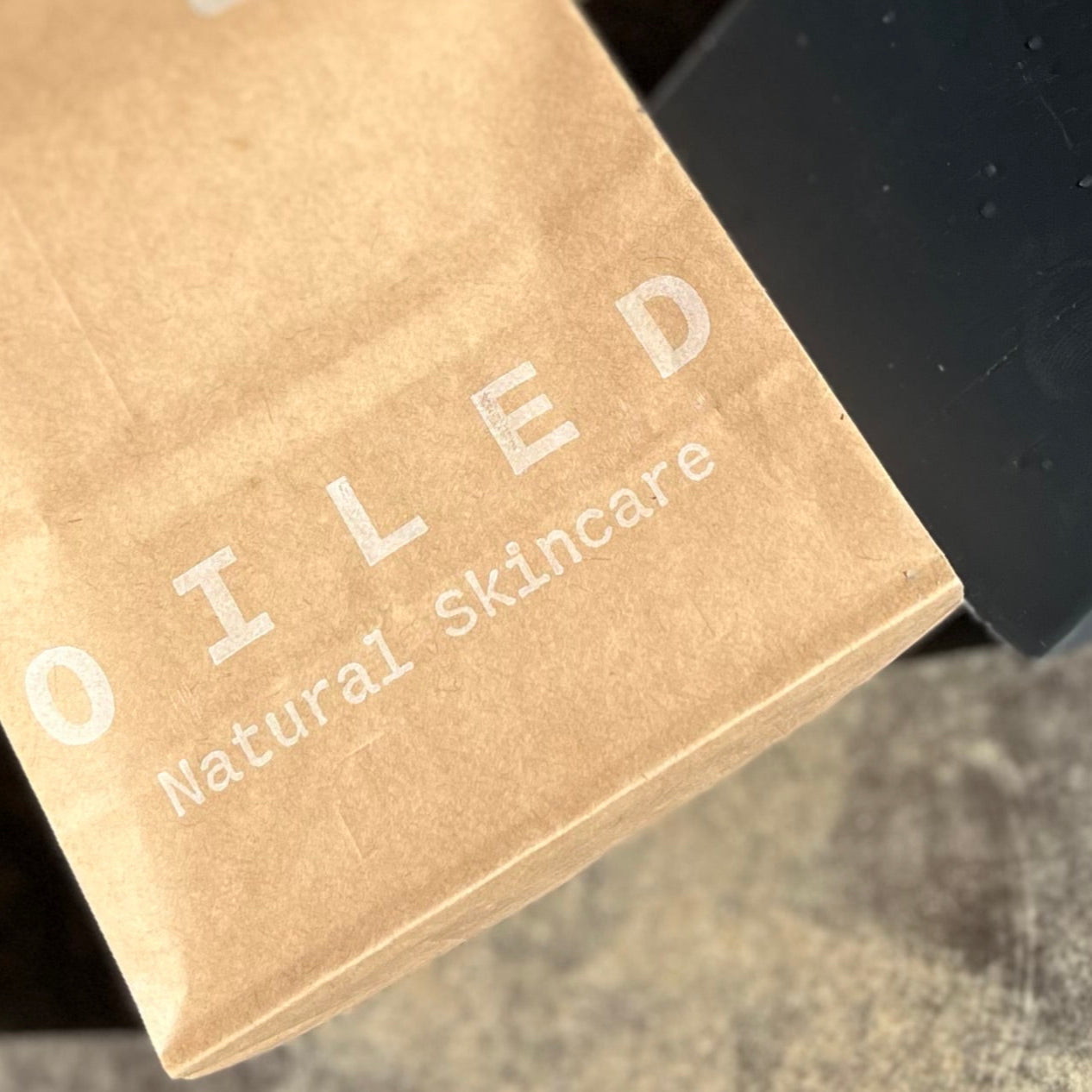 Brown paper bag printed with 'Oiled Natural Skincare' next to a black bar of soap.